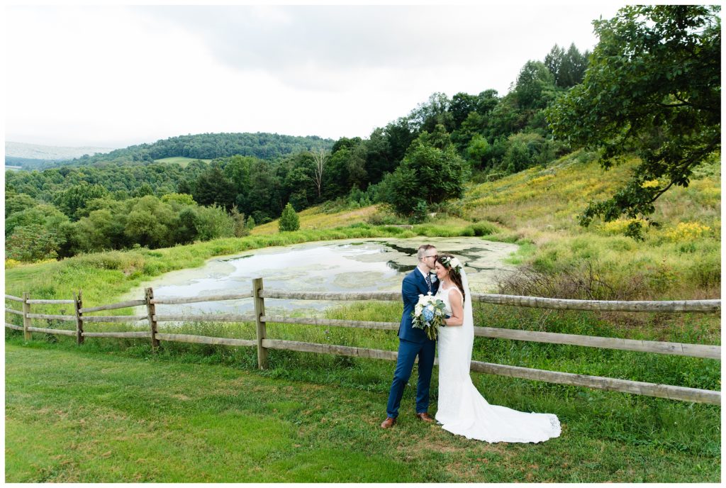 groom kissing bride's temple in front of wooden fence by pond on the mountainside