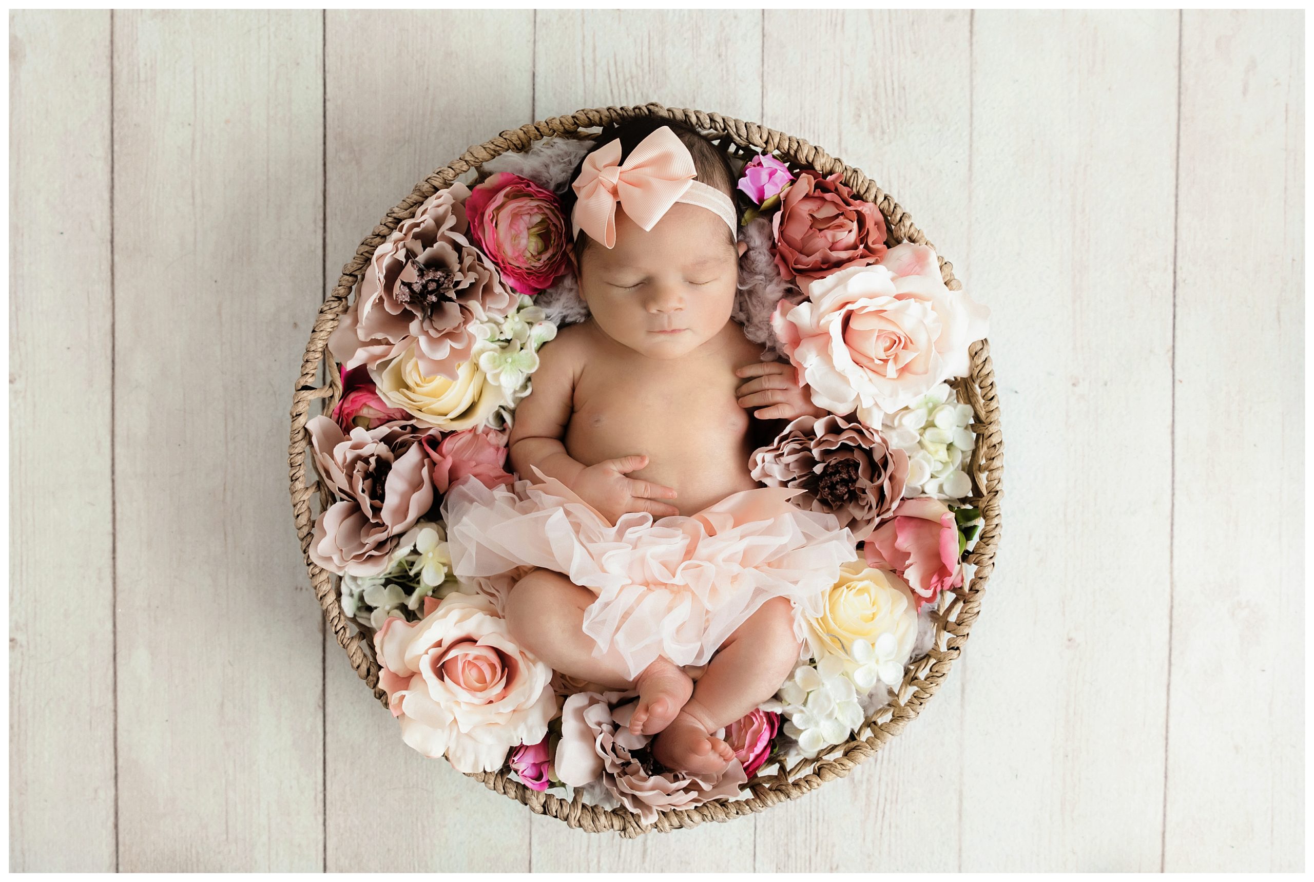 newborn girl in pink tutu and bow in basket surrounded by pink and cream flowers