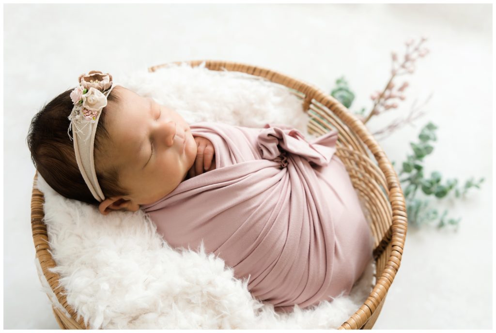 newborn girl wrapped in pink in basket with greenery