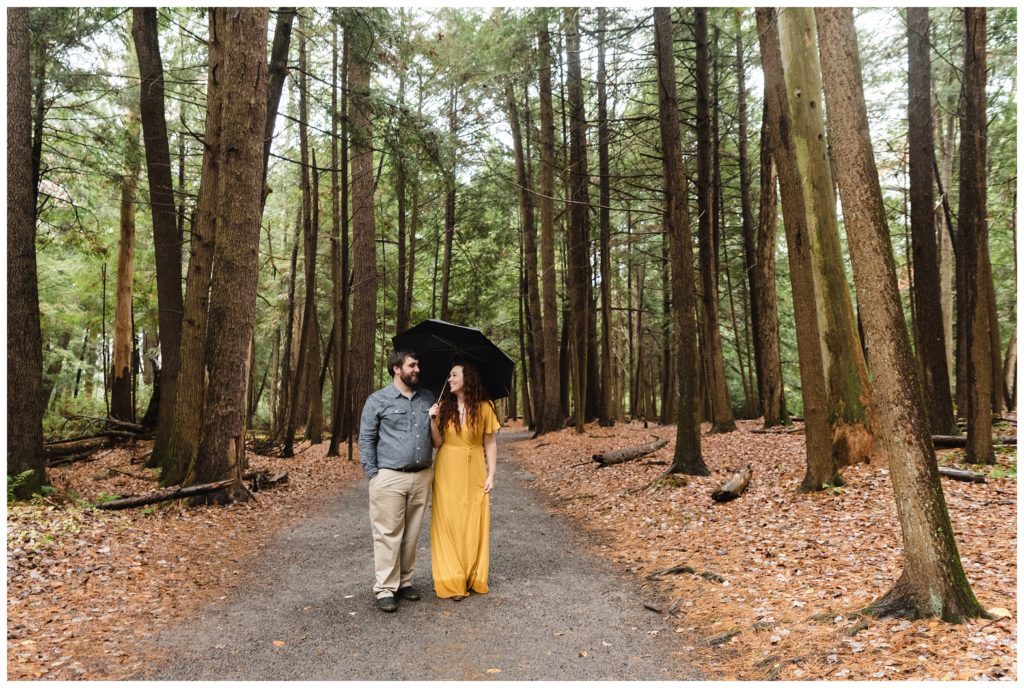 engaged couple walks along path at swallow falls holding umbrella and laughing with each other