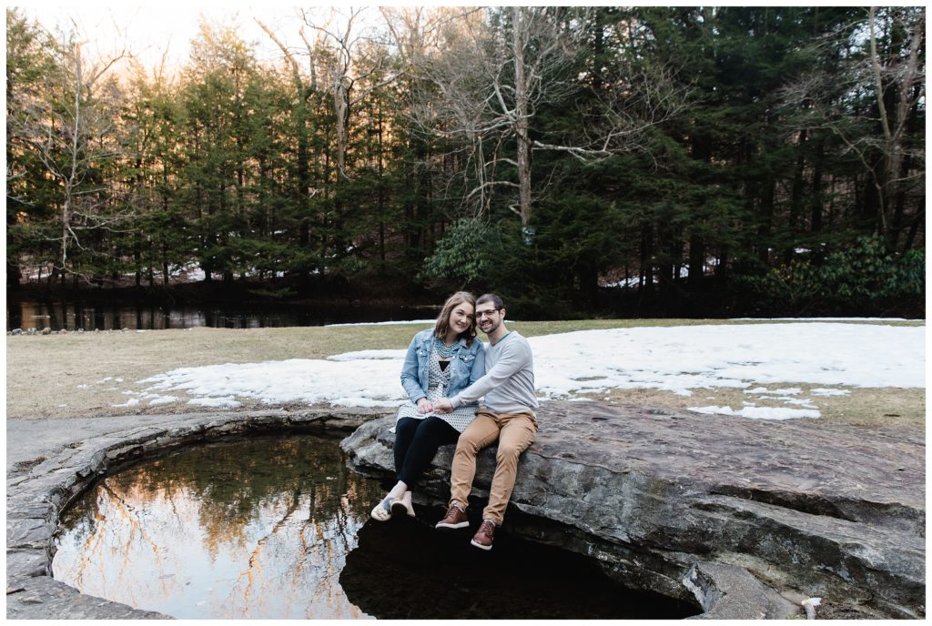 engaged couple sitting on rock over small pond