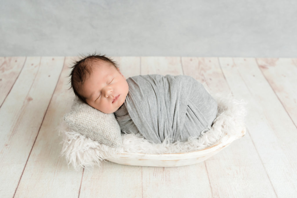 Baby boy in gray wrap sleeping on a pillow in a white bowl.