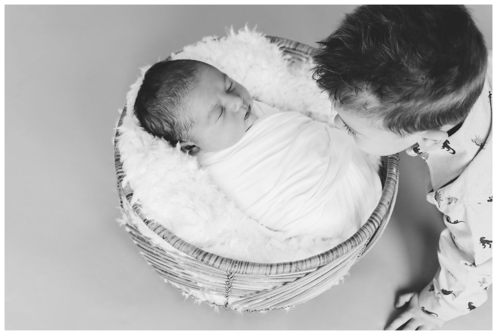 Black and white image of big brother looking down on newborn brother in basket.