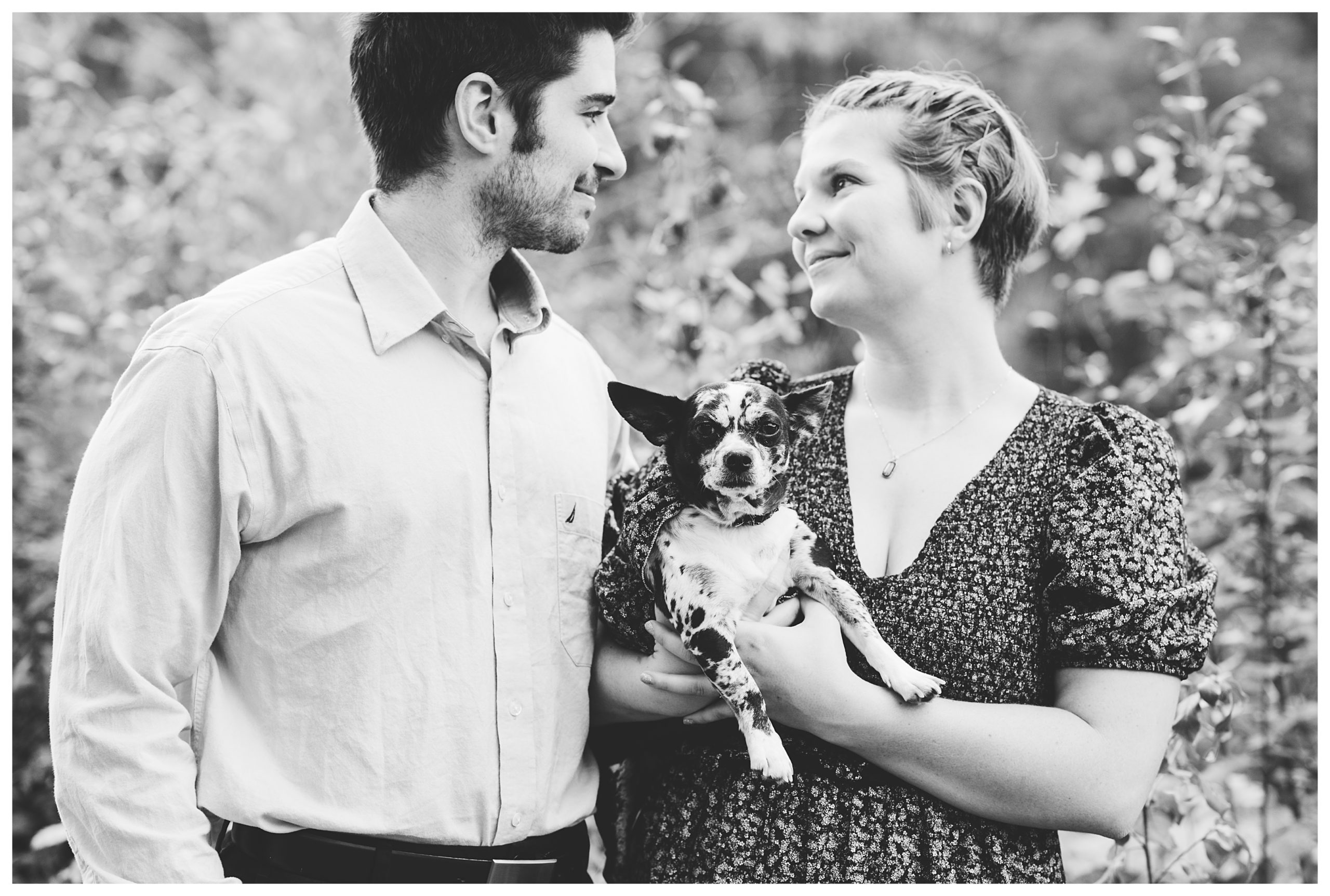 black and white of engaged couple looking at each other while holding their cute dog who looks directly at the camera
