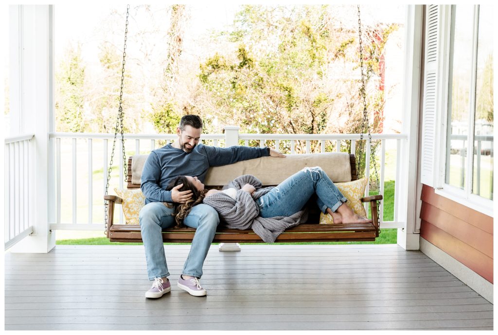 man holds fiancé's head in lap while she lies on porch swing- cozy home engagement session