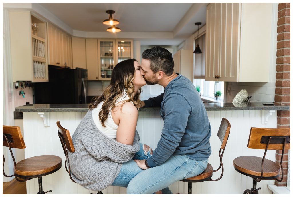 engaged coupe kiss on stools in front of their kitchen peninsula