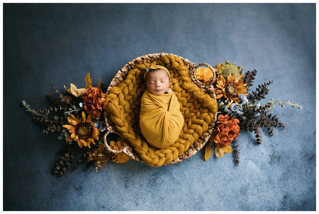 Newborn baby sleeping in yellow wrap on round basket, surrounded by fall blue and yellow flowers during pittsburgh studio newborn session