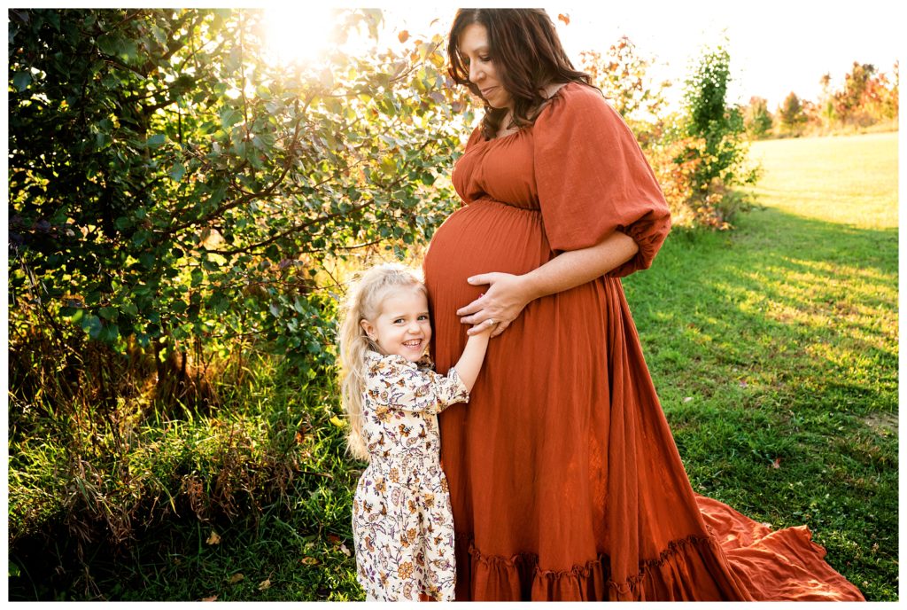 Pregnant mother wears rust maternity dress while daughter lovingly touches mom's belly