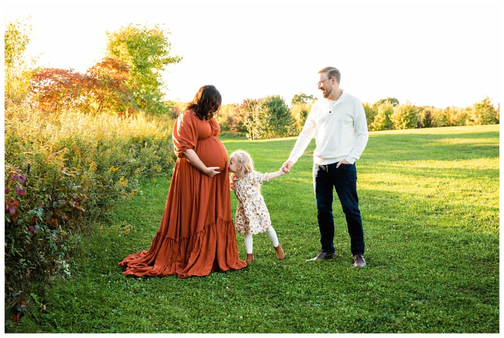 Family holds hands while daughter kisses mom's pregnant belly during maternity session in Bridgeville, PA.