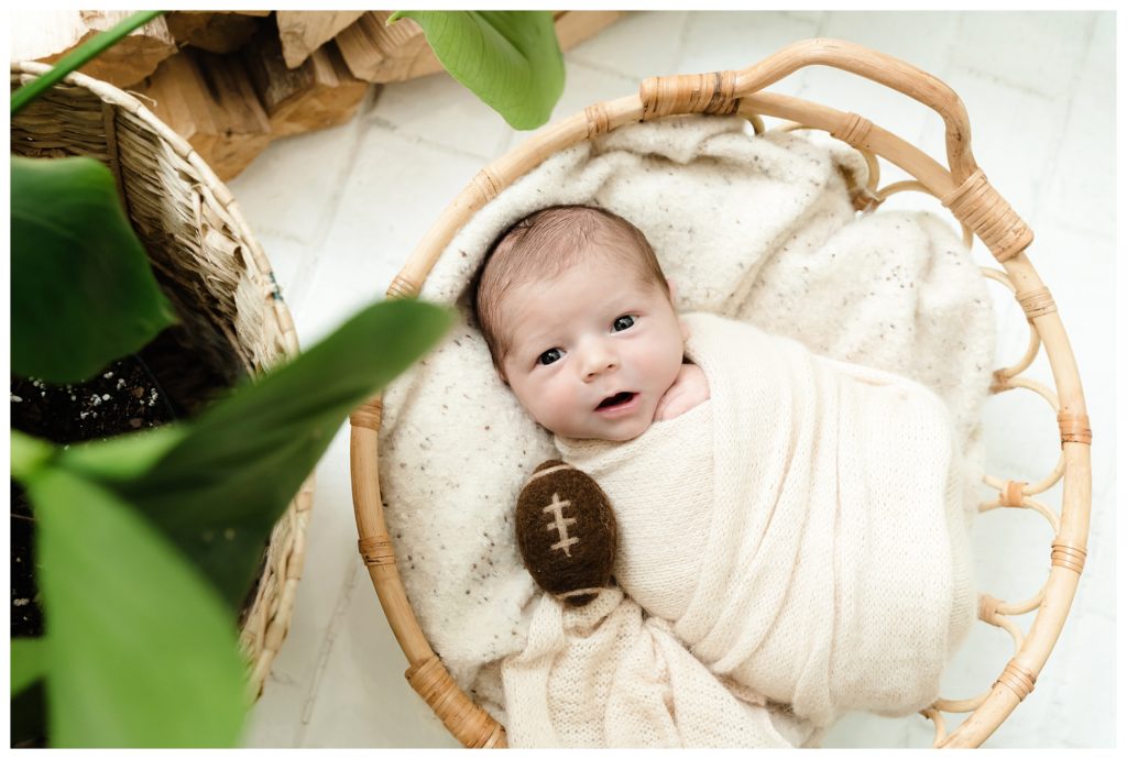 Baby Hudson Trubisky lays swaddled in basket with mini football during at-home lifestyle newborn session in Pittsburgh, PA.