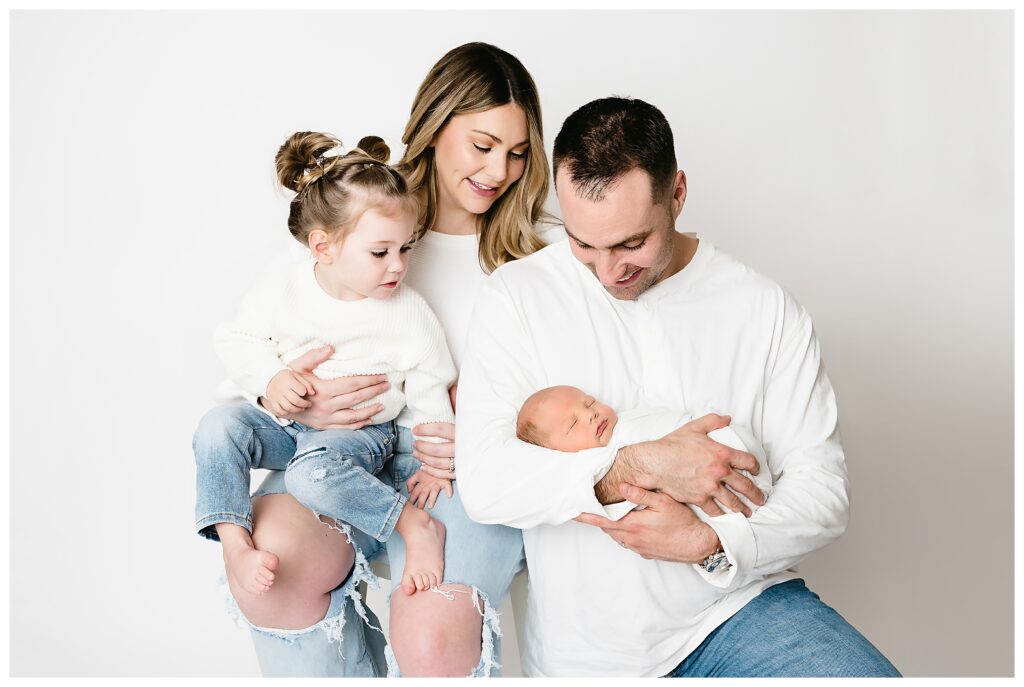 Mom, dad and big sister stare lovingly at newborn baby boy that dad is holding during their studio newborn session.