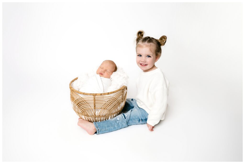 Big sister sits next to baby brother in boho basket on white backdrop  during their studio newborn session.