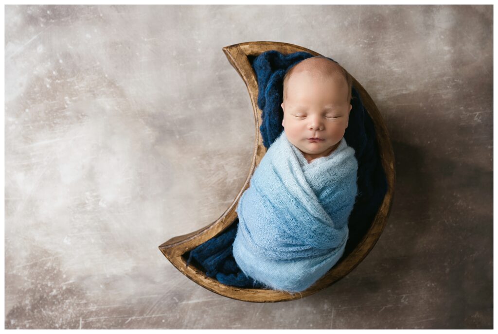 Newborn boy is swaddled in ombre blue wrap and sleeping inside wooden moon prop during his studio newborn session.