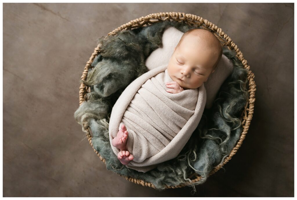 Newborn boy is wrapped in a tan swaddled with his cute toes showing while sleeping in a basket during his studio newborn session.