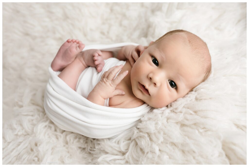Newborn baby boy looks at camera while swaddled in white on white flokati during his studio newborn session.