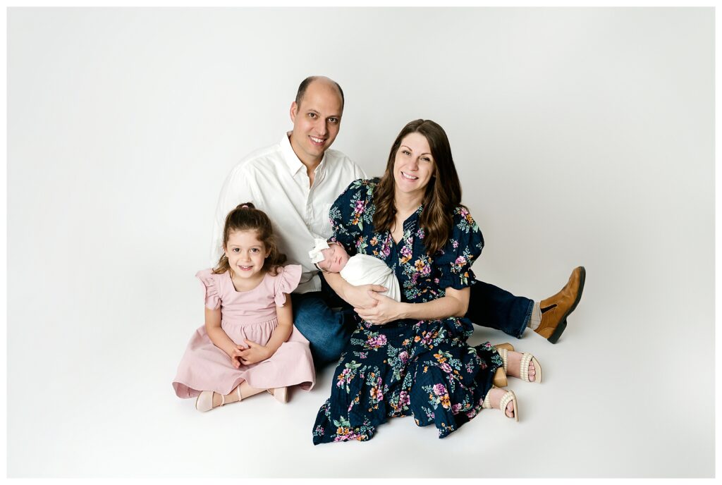 Family posed on floor of white backdrop holding newborn baby girl during Pittsburgh studio session.