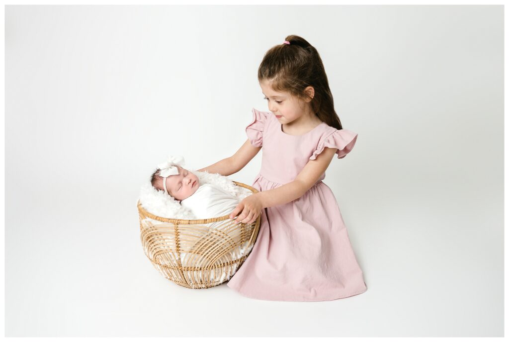 Big sister in pink dress kneels next to boho basket holding sleeping baby sister wrapped in white during Pittsburgh studio session.
