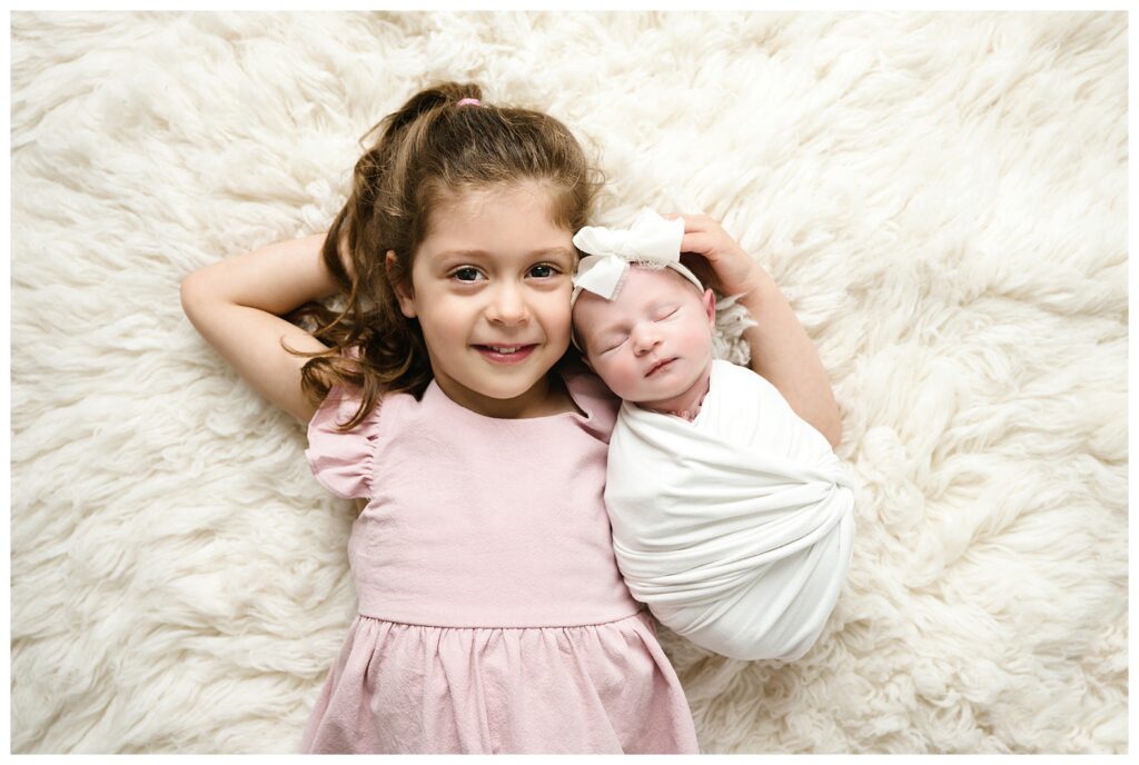 Big sister in pink dress lays on back while holding baby sister wrapped in white during Pittsburgh studio session.