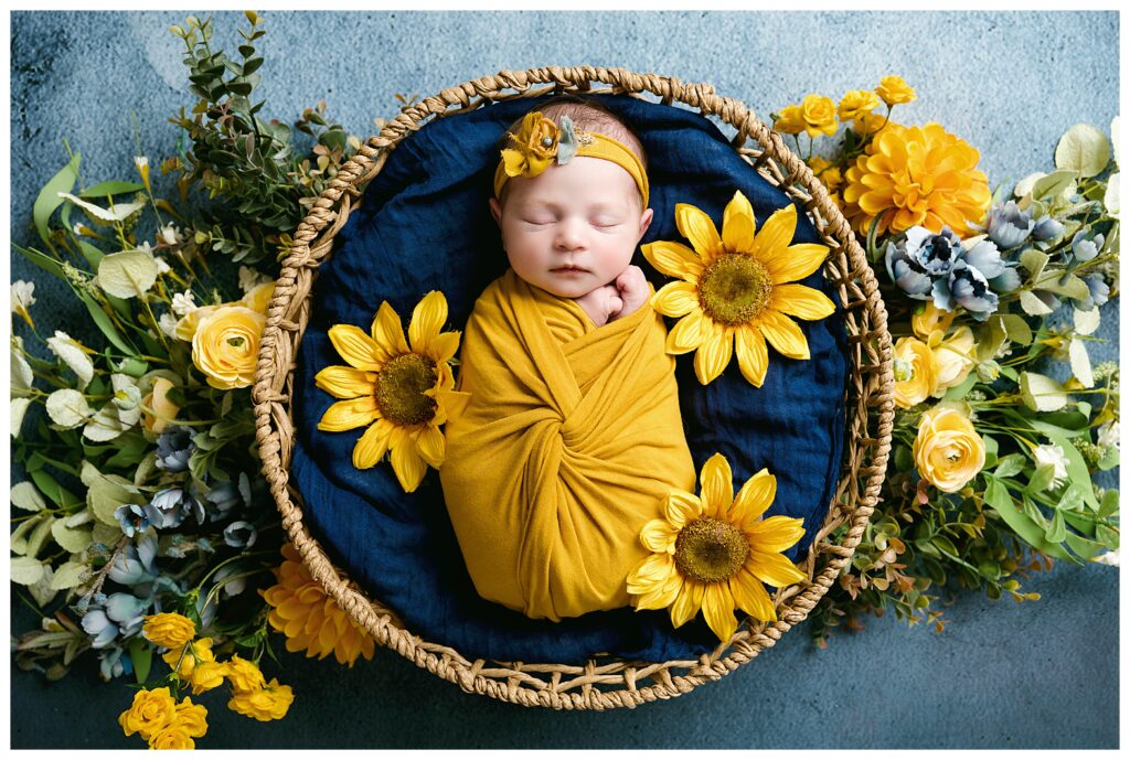 Newborn girl wrapped in mustard, sleeping in round basket on blue backdrop with sunflowers all around.