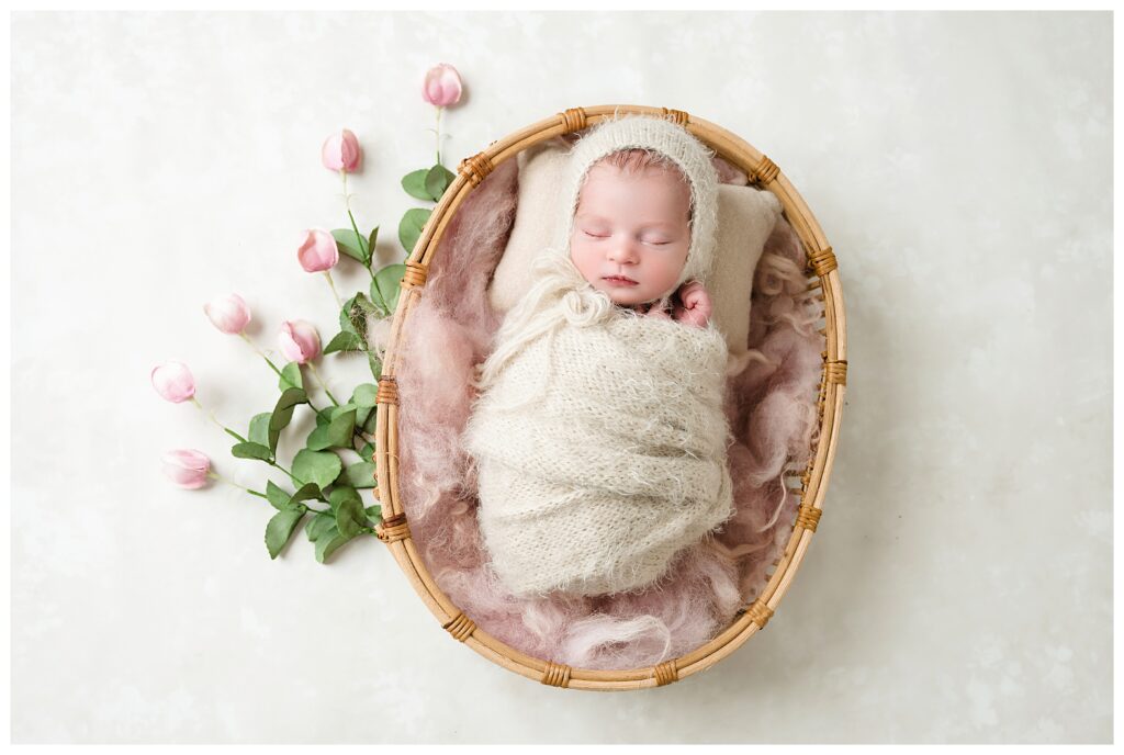 Baby girl wrapped in fuzzy neutral wrap with matching bonnet, sleeps on pink blanket in oval boho basket. Pink tulips lay beside the basket.