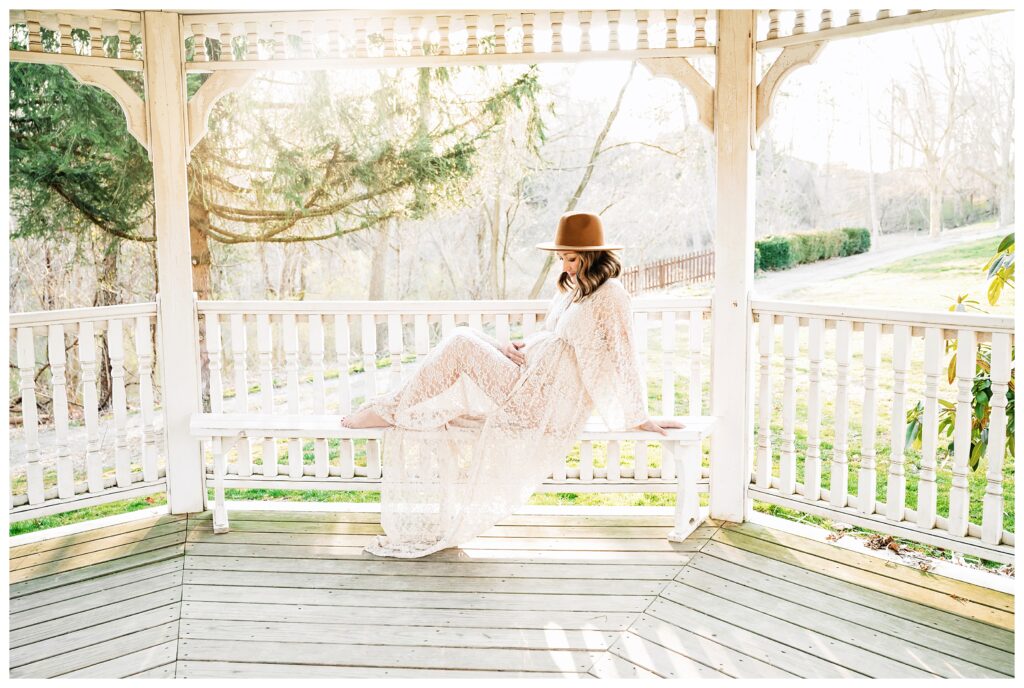 Pregnant mom wears lace dress and tan hat while sitting on a bench in the white gazebo at Robin Hill Park. The sun setting illuminates her from behind.