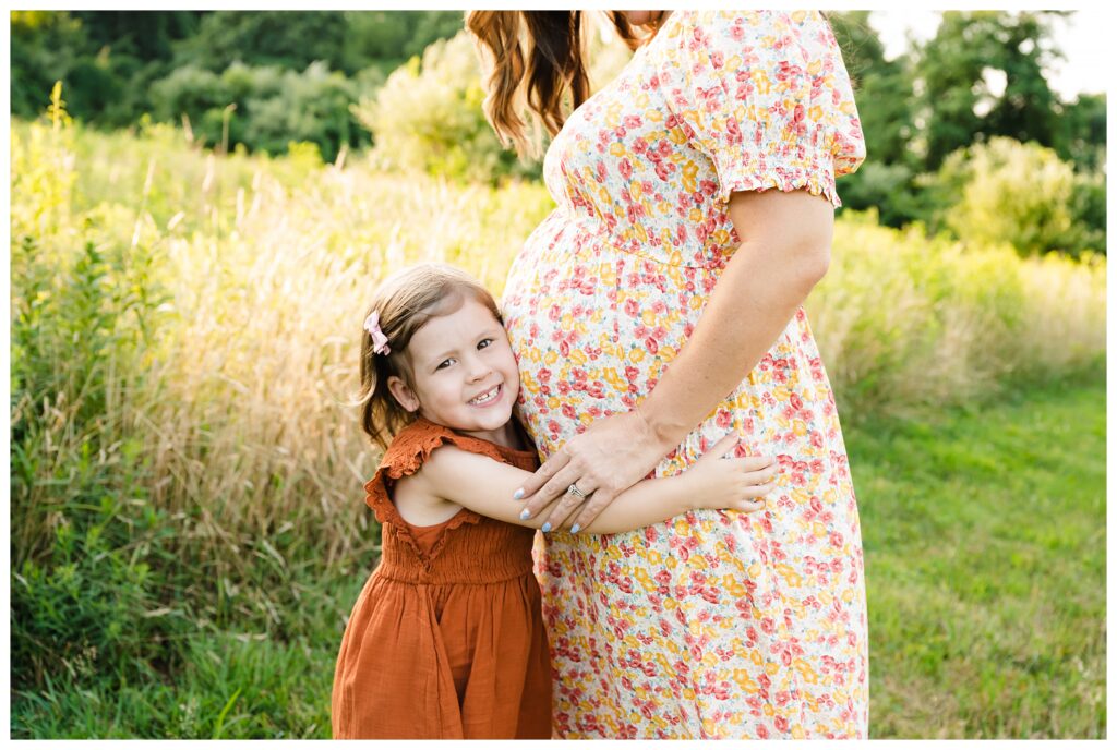 Young daughter embraces mom's pregnant belly with big smile in field at Mingo Creek Park.