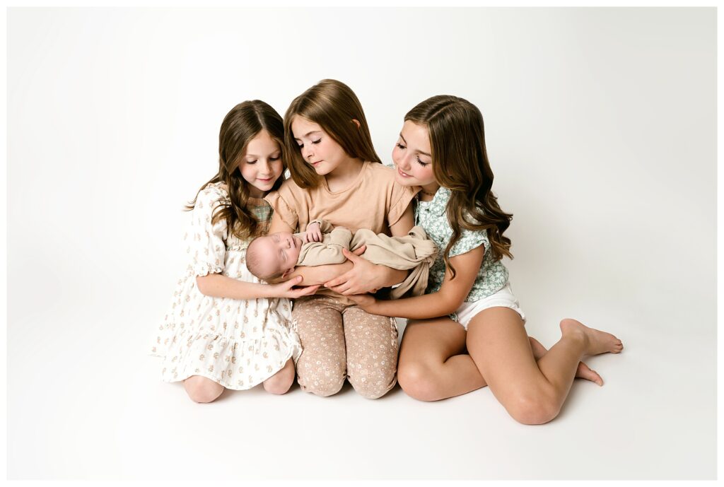 three older sister sit on needs on white backdrop while holding their newborn baby brother during this neutral studio newborn session.