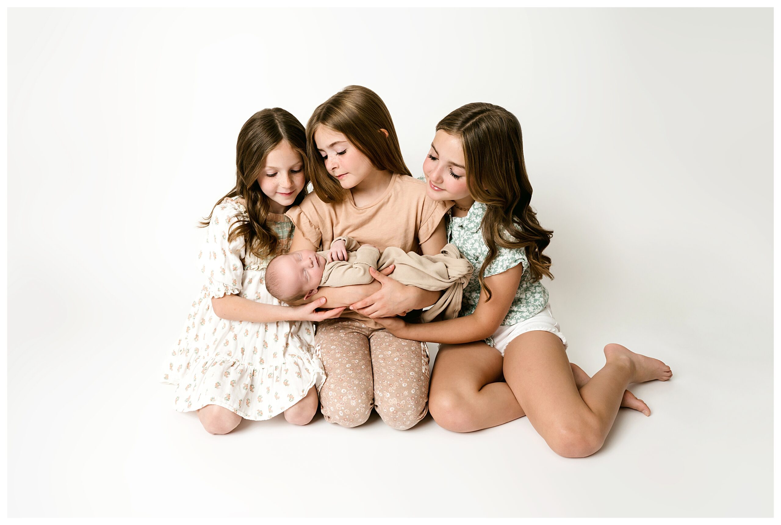three older sister sit on needs on white backdrop while holding their newborn baby brother during this neutral studio newborn session.