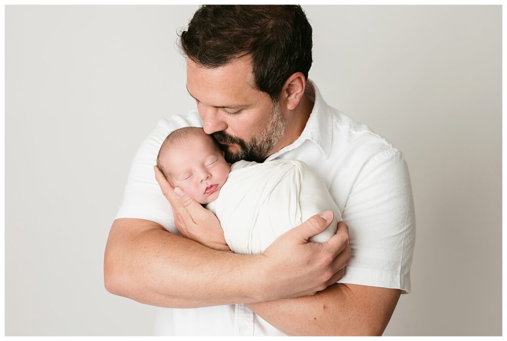 Dad snuggles newborn boy while kissing his temple during this neutral studio newborn session..