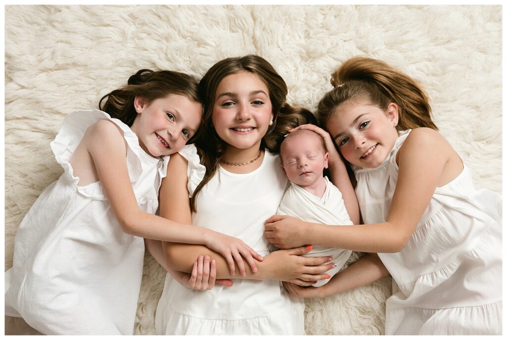 Three older sisters wear white and lay on backs looking up and smiling at the camera while holding their new baby brother.