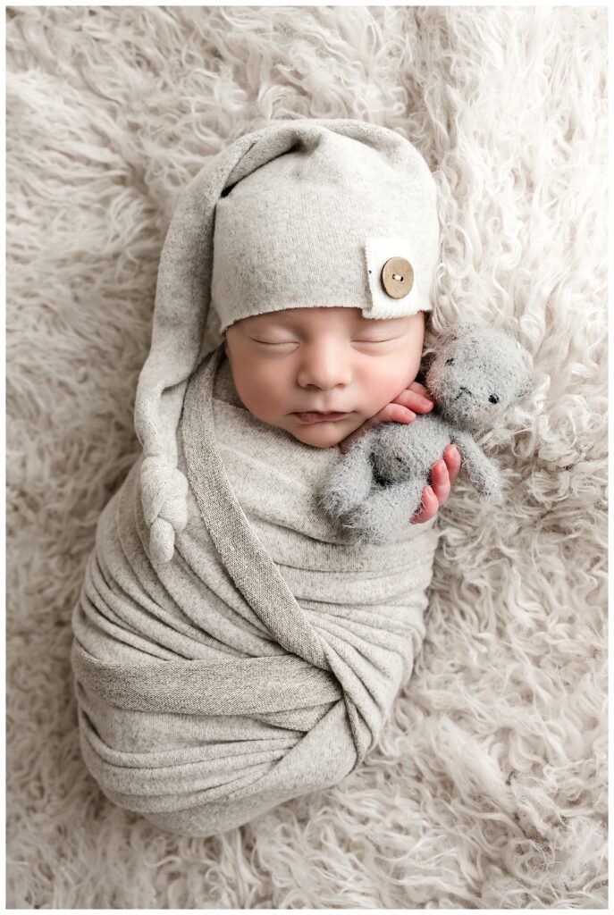 Newborn boy is wrapped in grey and wearing a cute sleepy cap while holding a grey teddy bear and sleeping during this neutral studio newborn session.