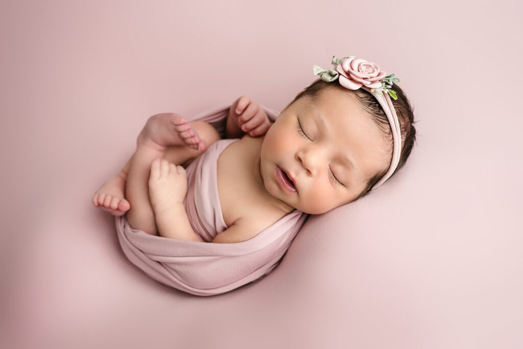 Newborn baby girls sleeps on a pink blanket with a pink wrap and a cute rose headband. Bridgeville newborn photographer.