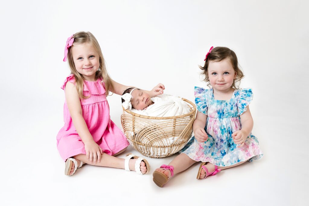 Two older toddler girls pose beside their baby sister who sleeps in a boho basket at Pittsburgh newborn studio. Bridgeville newborn photographer.