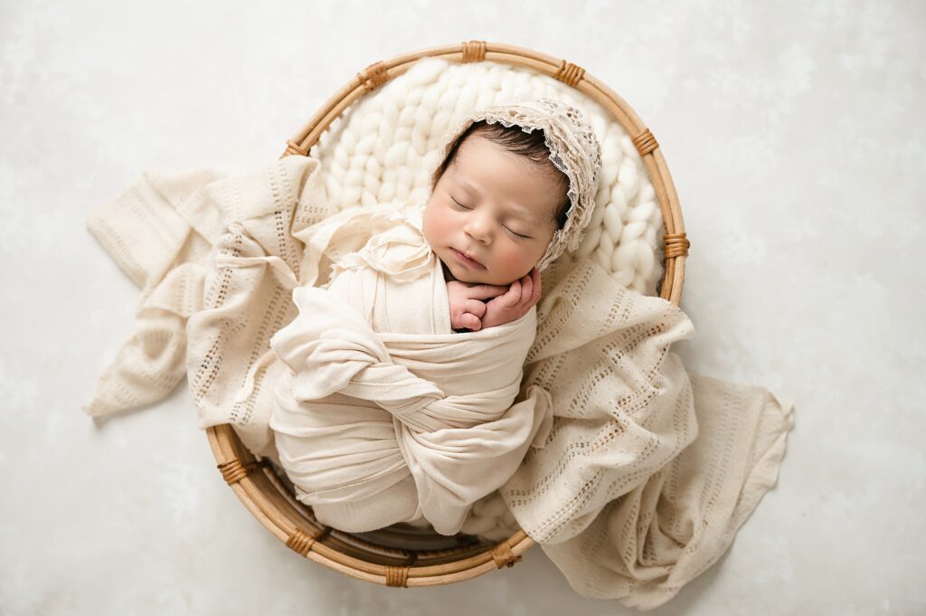 newborn baby girl posed in boho basket with cream blankets and wraps and a sweet lace bonnet