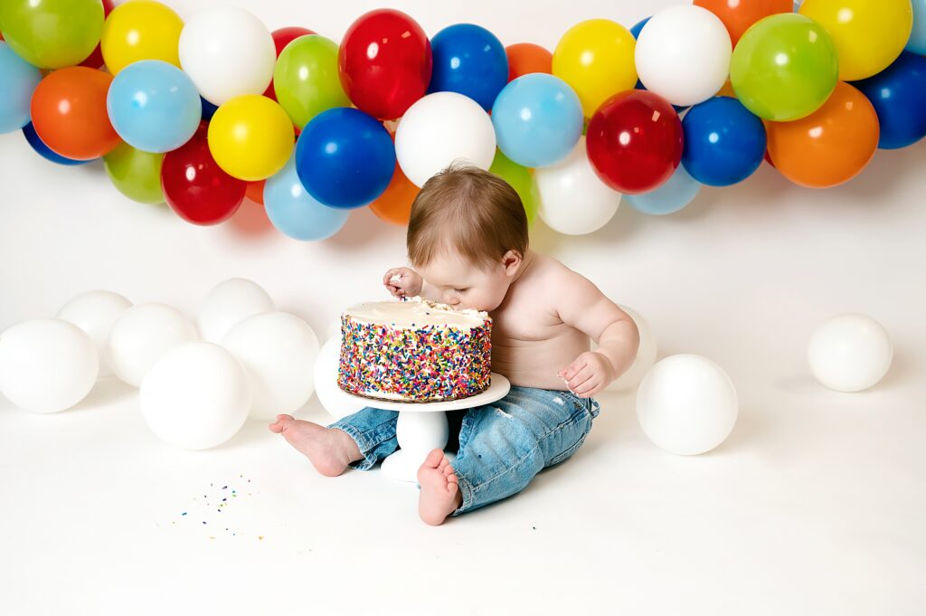 Baby boy tilts head to take a bite out of his cake with colorful balloons on a garland behind him baby during casual 1st birthday session.