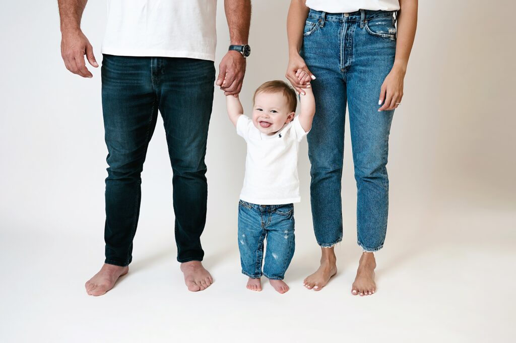 Cropped image of toddler standing in between parents legs, while reaching up and holding his hands baby during casual 1st birthday session.