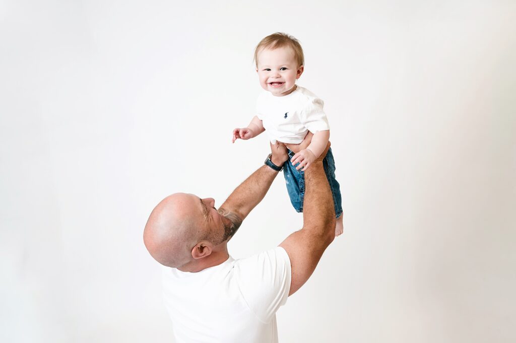 Dad lifting baby up into air while baby looks at camera smiling baby during casual 1st birthday session.