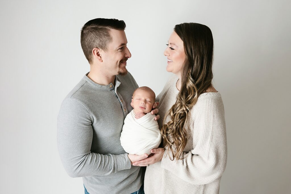 new parents hold newborn son swaddled between them as they look at each other and smile during pittsburgh baby photography session