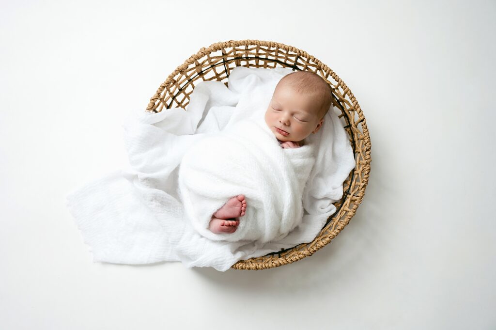 Newborn boy swaddles in white and sleeping in a round basket during pittsburgh baby photography session.