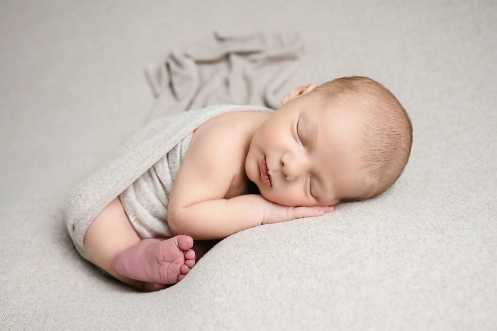 Newborn boy sleeps in taco position on tan backdrop during pittsburgh baby photography session.