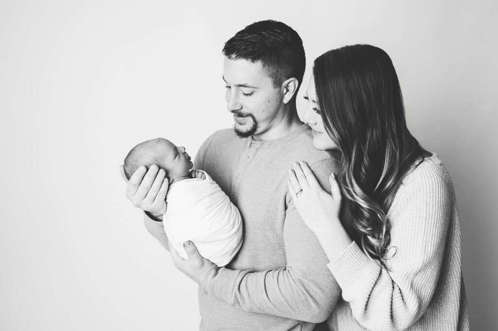 black and white image of dad hold newborn boy and mom leaning over dad's should while they both look at their baby during pittsburgh baby photography session