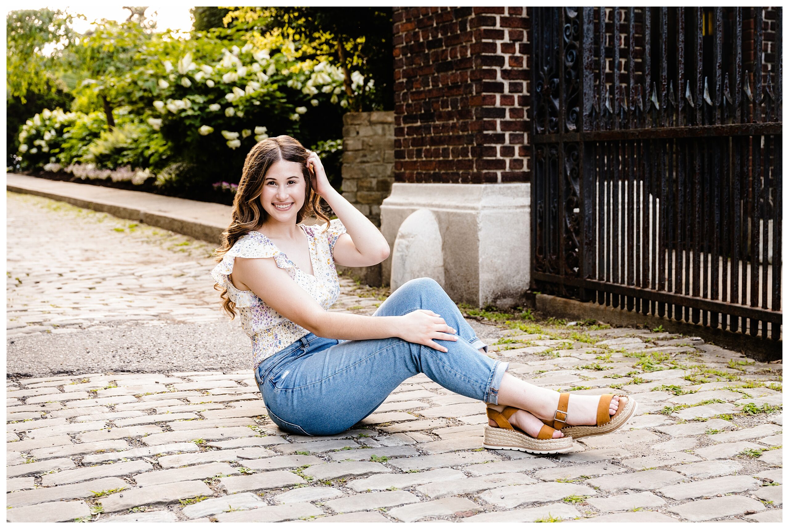 Senior girl sitting on a cobblestone street by the black iron gates at Mellon Park in Pittsburgh, smiling confidently in jeans and a floral top. Using Katie Louise Photography as her Pittsburgh senior photographer.