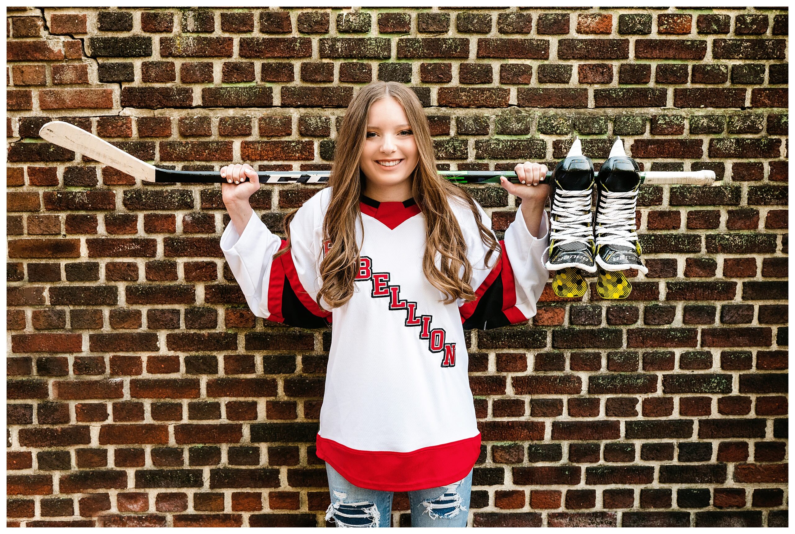 Senior girl wearing a Rebellion hockey jersey and holding a hockey stick over her shoulders with her skates hanging from it, posing in front of a brick wall of Mellon Garden, Pittsburgh.